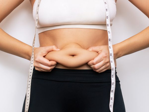 Cropped shot of a young woman 
with measuring tape around the neck holding herself by the fat on her stomach isolated on a white background. Overweight, excess weight concept